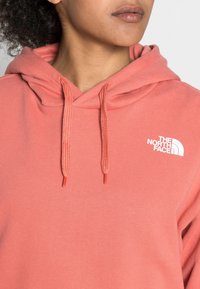 Koraalrode hoodie gemaakt van een katoenmix, met een capuchon met trekkoord, geribbelde manchetten en een wit logo op de borst. Soepele textuur, casual ontwerp.