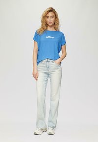 Blauwe t-shirt met korte mouwen en witte tekst "s.Oliver," gecombineerd met lichtgewassen jeans en lichte sneakers. Informele, relaxte pasvorm.