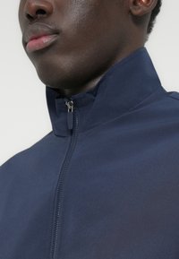 Κοντινό πλάνο στο κάτω μέρος του προσώπου και το λαιμό ενός ατόμου που φοράει μπλε σκούρο μπουφάν με φερμουάρ μέχρι ψηλά και όρθιο γιακά.