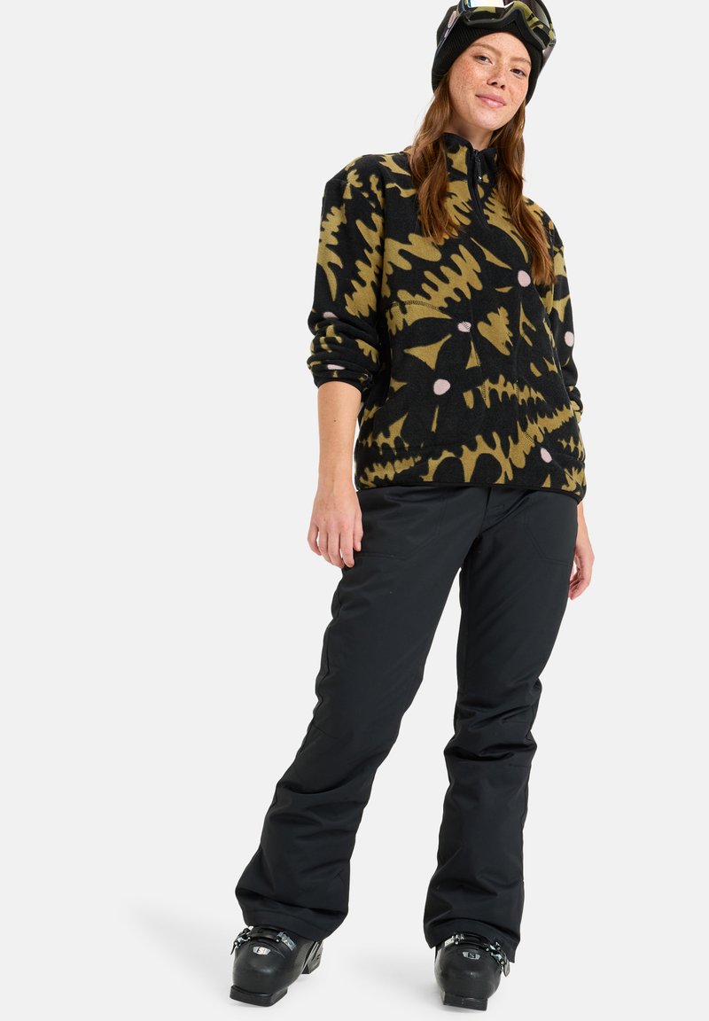 Pullover in pile nero con un audace motivo giallo e rosa, colletto con zip e pantaloni isolanti neri; abbinato a stivali da sci neri e a un berretto.