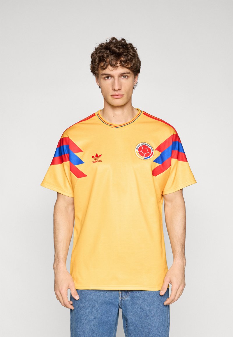 Maglia gialla a maniche corte con accenti grafici blu e rossi sulle spalle. Presenta uno scollo rotondo e un logo circolare sul petto.