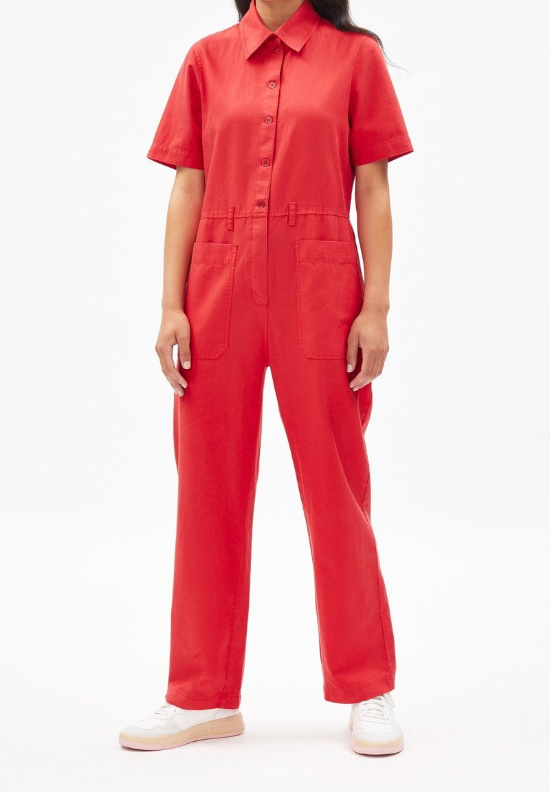Femme portant une combinaison rouge à manches courtes avec des poches à l'avant et des baskets blanches, debout devant un fond blanc.