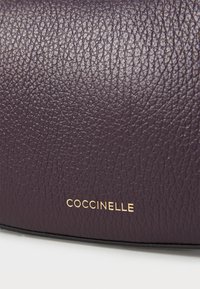 Tamsiai violetinė odinė rankinė su tekstūruotu paviršiumi ir auksiniu "Coccinelle" logotipu išpjaustytu apačioje.