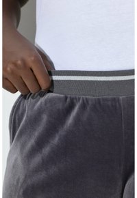Grau melierte Velours-Sweatpants mit weicher Textur, ausgestattet mit einem gerippten Bund mit zwei horizontalen weißen Streifen. Hand greift den Bund.