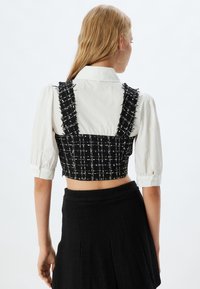 Zwart-wit geblokte cropped top met rafelige banden, gecombineerd met een witte blouse met kraag. Gelaagd stof met zichtbare stiksels.
