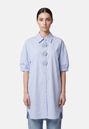 Donna che indossa una camicia oversize azzurro chiaro con tre grandi bottoni rotondi e maniche corte a sbuffo, abbinata a jeans blu su uno sfondo semplice.