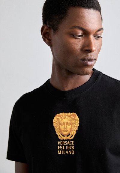 Schwarzes Baumwoll-T-Shirt mit grafischem Medusa-Motiv in Gold und dem Text "VERSACE EST. 1978 MILANO" in fetter Schrift nahe dem Rundhalsausschnitt.