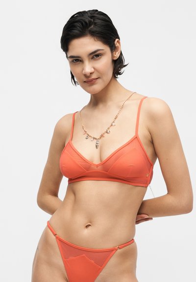Γυναίκα με κοντά σκούρα μαλλιά που φοράει ένα ταιριαστό πορτοκαλί σετ σουτιέν και σλιπ, ποζάροντας με τα χέρια της πίσω από την πλάτη της σε ανοιχτό φόντο.