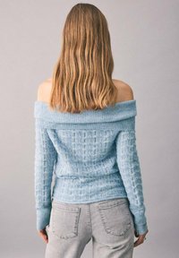 Femeie cu păr ondulat de lungime medie, purtând un pulover tricotat bleu deschis fără umeri și blugi gri deschis, stând în fața unui fundal simplu.