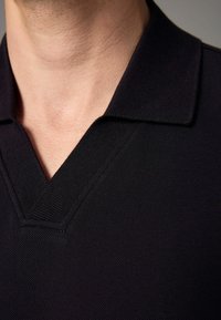 Svart pikétröja med strukturstickat tyg, med en framträdande V-ringning och platt krage. Enfärgad design, utan synliga mönster eller detaljer.