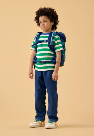  camiseta de rayas verde y blanca, mochila azul, jeans azul, y zapatillas blancas con acentos verdes y amarillos. De pie sobre un fondo claro.