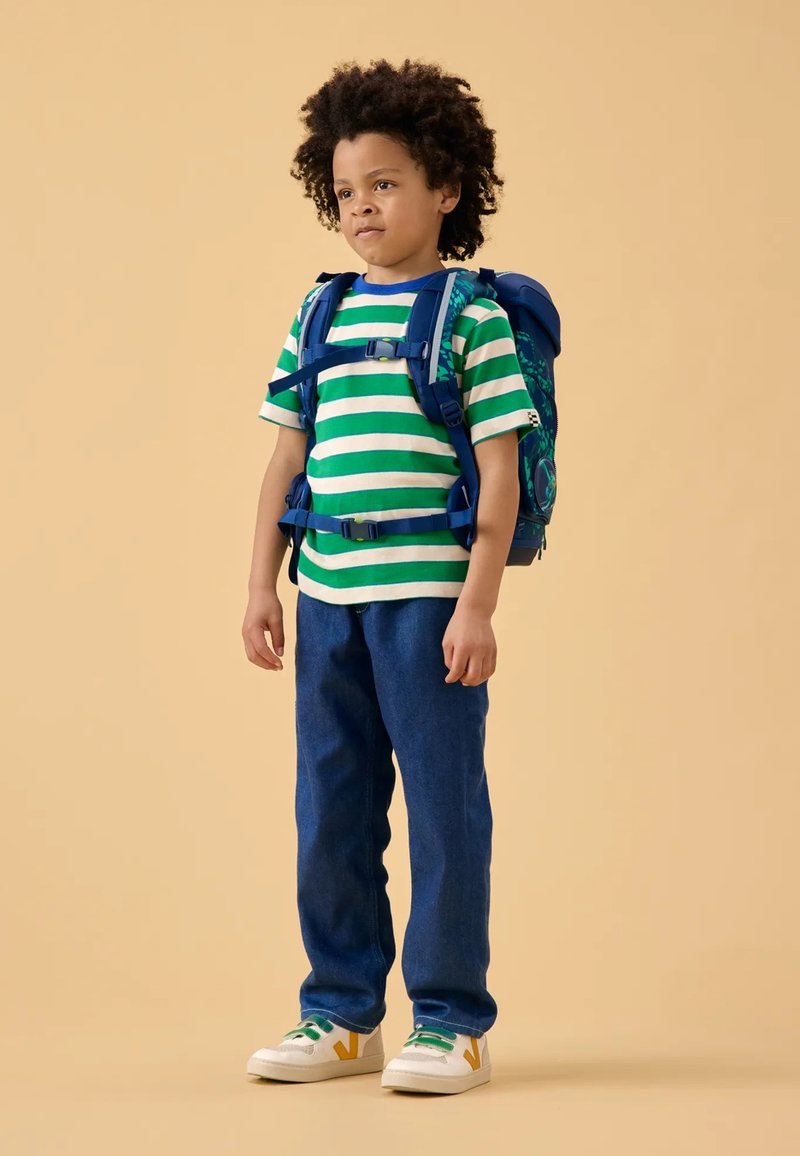 Grün-weiß gestreiftes T-Shirt, blauer Rucksack, blaue Jeans und weiße Turnschuhe mit grünen und gelben Akzenten. Auf einem hellen Hintergrund stehend.