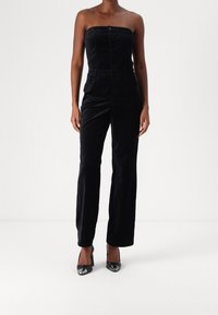 Svart axelbandslös jumpsuit i mjuk manchester med en figurnära bodice, raka vida ben och framknäppning. Matchas med blanka svarta klackar.