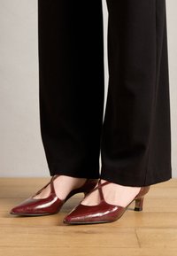Chaussures en cuir verni bourgogne avec un bout pointu, un design découpé et un petit talon bloc, portées avec un pantalon noir sur un sol en bois.