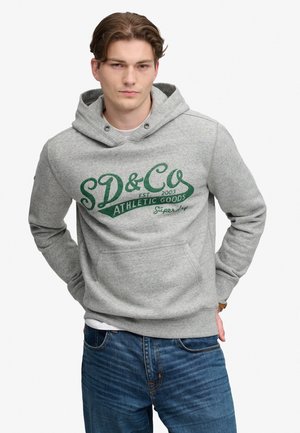Jonge man met kort bruin haar, gekleed in een grijze hoodie met groene "SD&Co Athletic Goods" tekst en blauwe spijkerbroek, staand met handen achter de rug.