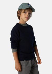 Suéter azul marino de mangas largas, cuello redondo; camiseta de mangas largas gris debajo. Gorra gris con logo, pantalones cortos gris claro. Persona joven posando.
