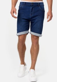 Shorts en denim bleu foncé, avec des ourlets roulés et une fermeture à bouton, associés à des baskets blanches. Tissu texturé, design décontracté.