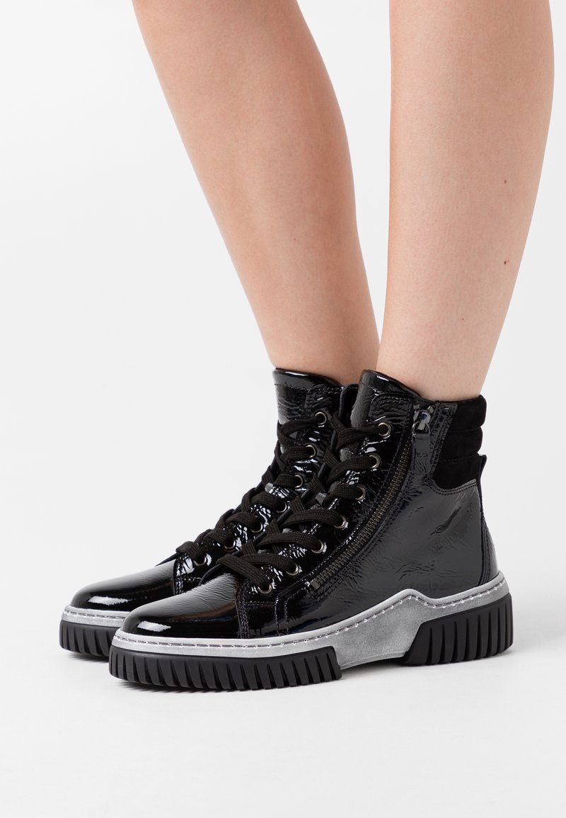 Botas altas de charol negro con cordones negros, cremalleras laterales y suelas de goma gruesa en negro y gris con tacos, luciendo piernas al aire.
