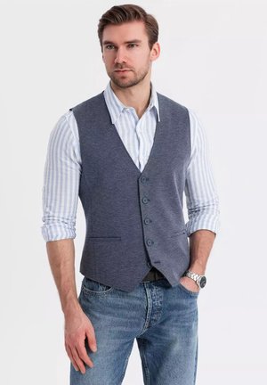 Gilet en tricot gris avec cinq boutons, porté sur une chemise rayée bleu clair. Associé à un jean bleu et une montre argentée. Design simple.