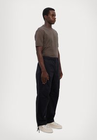 Camiseta de algodão marrom com gráfico circular; calças cargo pretas com múltipos bolsos; ténis brancos com sola texturizada.