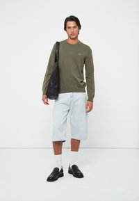 Olijfgroen lange mouwen trui, lichtblauwe denim shorts tot de knie, witte sokken, zwarte instap schoenen en een zwarte schoudertas.