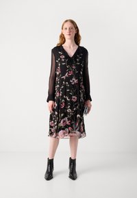 Schwarzes Blumenkleid mit langen durchsichtigen Ärmeln, V-Ausschnitt und Rüschendetails. Hat rosa und grüne Blumenmuster. Kombiniert mit schwarzen Ankle Boots.