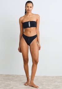 Juodas straples bikinio viršus su baltu logotipu; derantis aukšto liemens bikinio apačia. Lygi medžiaga, minimalios apimties dizainas. Nėra ryškių raštų.
