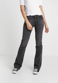 Jean noir évasé en tissu denim souple, avec une taille mi-haute, un design à cinq poches et des passants pour ceinture, porté avec des baskets noires.