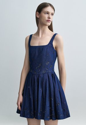 Žena v tmavomodrých šatách bez rukávov so štvorcovým výstrihom a rozptýlenými malými kvetinovými ozdobami, ktorá hľadí doprava na jednotnom pozadí.