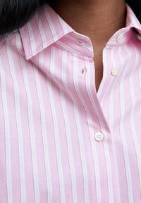 Femme portant une chemise à boutons rayée verticalement rose et blanche, col ouvert, montrant le cou et la partie supérieure de la poitrine.