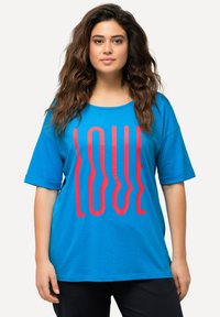 Camiseta azul de manga corta con un gran gráfico en rojo ondulado que dice "LOVE". Cuello redondo, corte relajado y textura suave. Combinada con pantalones negros.