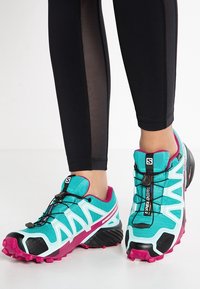 Türkise und schwarze Sportschuhe mit pinken Akzenten, ausgestattet mit einem strukturierten Obermaterial und aggressivem Profil für guten Halt, sowie einem Schnürsystem für einen sicheren Sitz.