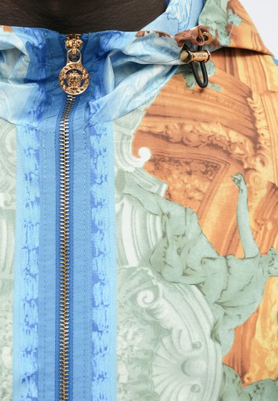 Barvna vetrovka z zadrgo, okrašena z izpopolnjenimi baročnimi vzorci v oranžni in svetlo zeleni barvi, z modrim teksturiranim panelom in z zlatimi dodatki.