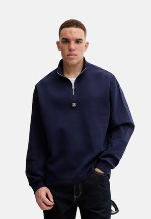 Sudadera con cremallera en color navy, de corte relajado, con un cuello alto y un pequeño logo en el pecho. Tejido texturizado y puños acanalados.