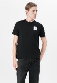 T-shirt noir en coton avec un col rond et un petit écusson blanc CK sur la poitrine gauche ; assorti à un jean foncé.
