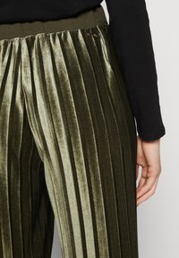 Pantaloni plissettati in velluto verde oliva scuro, con una vita spessa e una leggera lucentezza, e un accento del marchio ben visibile sul lato.
