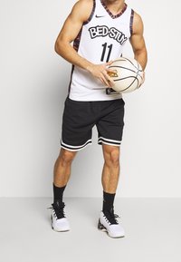 Maillot de basket blanc avec bordures multicolores, affichant "BED-STUY" et le numéro "11". Short noir à rayures blanches, chaussures noires, tenant un ballon de basket.