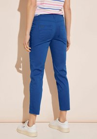 Blue, fitted trousers with a smooth texture, cropped length, and back pockets. Paired with white sneakers against a light background.