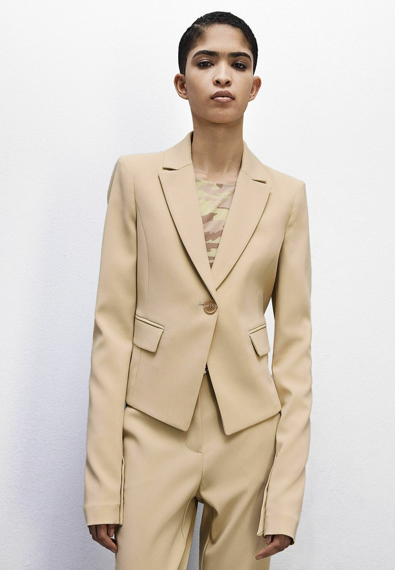 Blazer beige su misura con un bottone, rever a punta e tasche frontali; indossato sopra un top con motivo mimetico, caratterizzato da tessuto morbido.