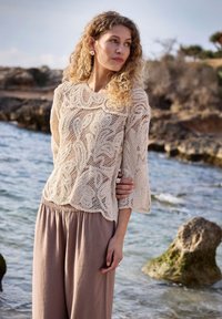 Femme aux cheveux bouclés portant un pull en dentelle beige et un pantalon taupe ample, debout près d'un rivage rocheux avec une mer calme en arrière-plan.