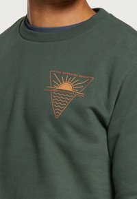 Grön sweatshirt med rund hals, prydd med en triangelformad, orange grafisk design av en sol och vågor, tillverkad av bomull eller liknande material.