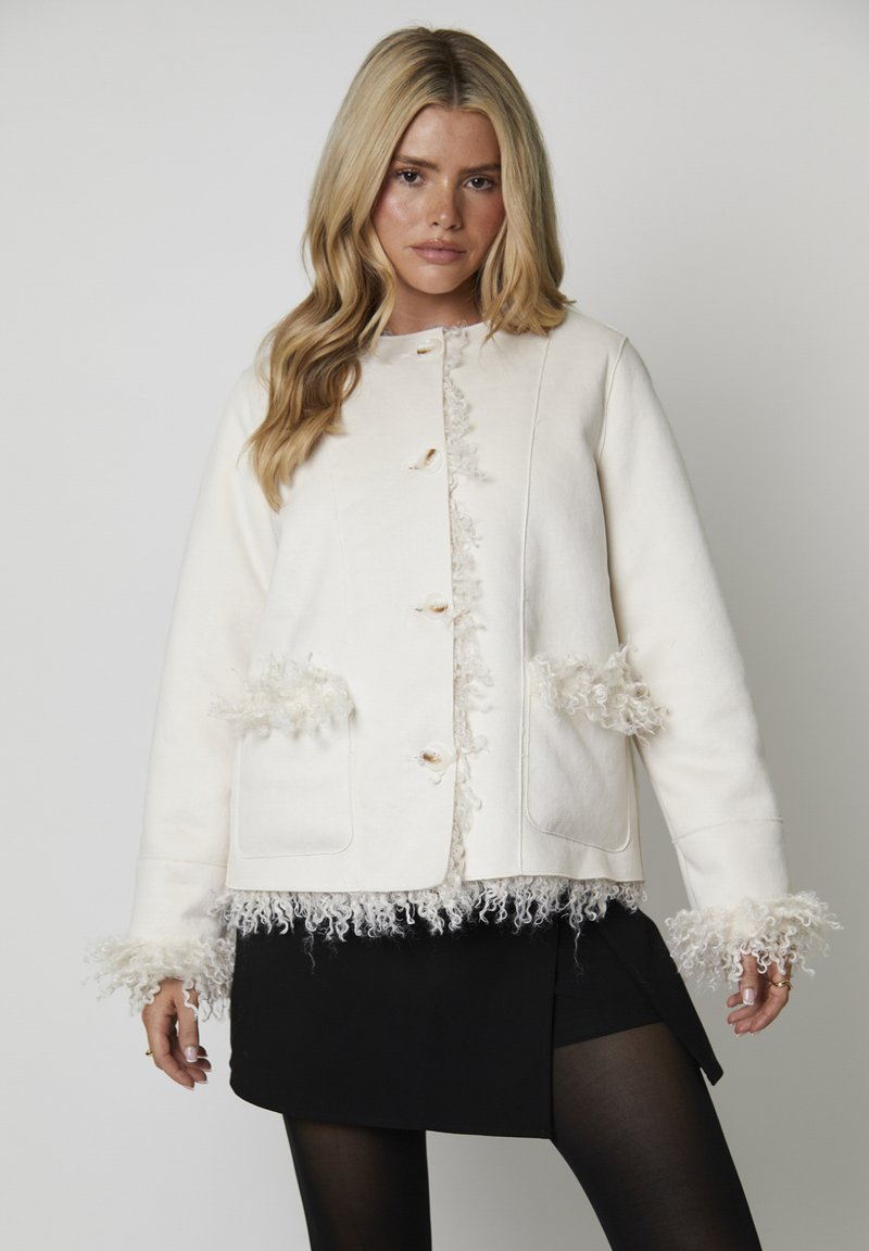 Biała kurtka z teksturowanym frędzlem na kieszeniach i mankietach, o prostym kroju i okrągłym dekolcie. Guziki dodają detalu.