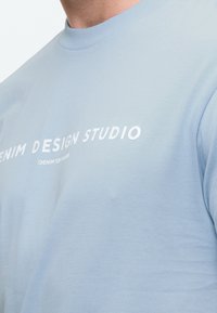 Γαλάζιο βαμβακερό T-shirt με στρογγυλή λαιμόκοψη, με άσπρο κείμενο "DENIM DESIGN STUDIO" τυπωμένο στο στήθος. Λείος υφή.