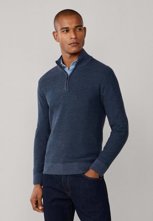 Suéter de cremallera azul marino hecho de tejido de punto texturizado, con un cuello alto y puños acanalados. Llevado sobre una camisa de cuello azul claro.
