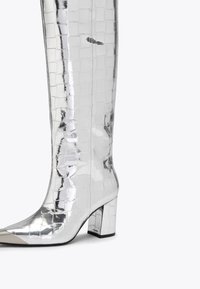 Srebrne wysokie botki do kolan o błyszczącej powierzchni, z teksturowanym kwadratowym wzorem, spiczastym noskiem i słupkowym obcasem.