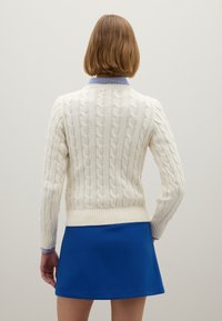 Frau mit kurzen braunen Haaren, die einen weißen Pullover mit Zopfmuster, einen Kragen mit blauen Streifen und einen blauen Rock trägt, steht vor einem schlichten Hintergrund.