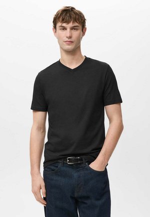 Jonge man met kort lichtbruin haar, draagt een zwart V-hals T-shirt en donkere spijkerbroek met een zwarte riem, staand met één hand in de zak.