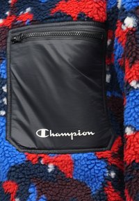 Black zippered pocket on a colorful fleece garment; features red, blue, and navy patterns; prominent Champion logo in white.