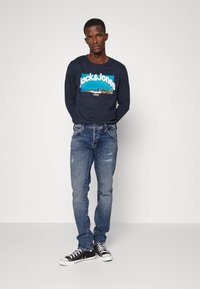 Camisa de manga larga navy con gráfico de "Jack & Jones"; combinada con jeans claros desgastados y zapatillas negras. Diseño ajustado y casual.