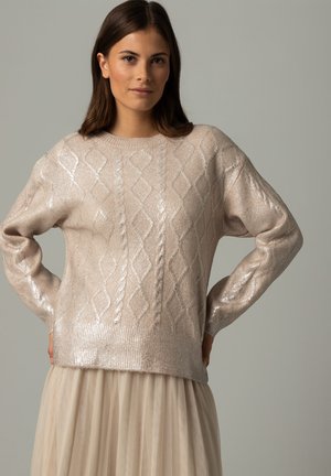 Pull beige clair en tricot doux et brillant avec des motifs en losange et des poignets côtelés. Accordé avec une jupe fluide de couleur neutre.